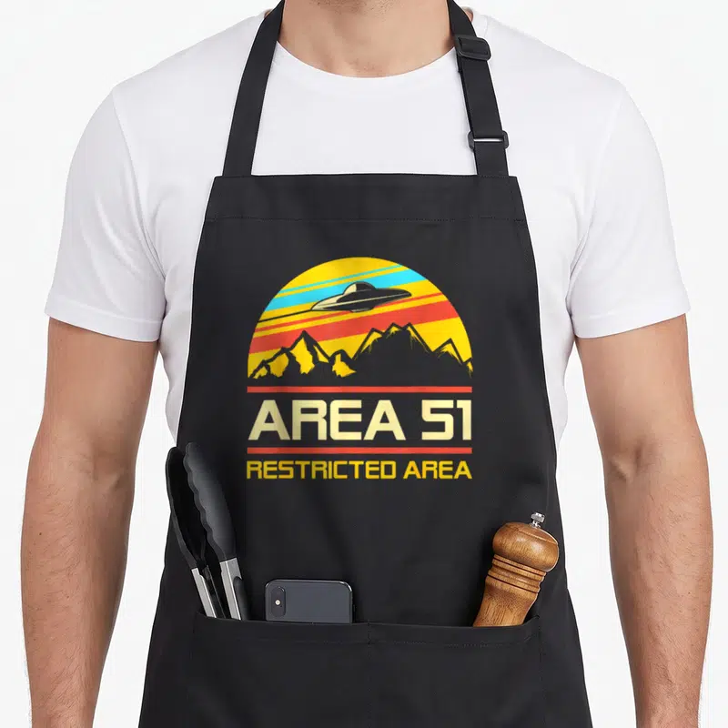 Tablier de cuisine noir avec logo Area 51 rétro montrant un vaisseau spatial au-dessus de montagnes aux couleurs vives.