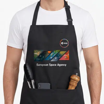 Tablier de cuisine noir avec le logo ESA et une illustration diagonale représentant des planètes, des fusées et des stations orbitales.