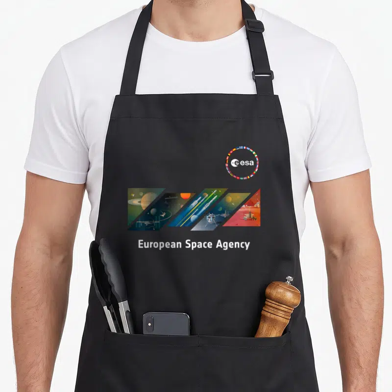 Tablier de cuisine noir avec le logo ESA et une illustration diagonale représentant des planètes, des fusées et des stations orbitales.