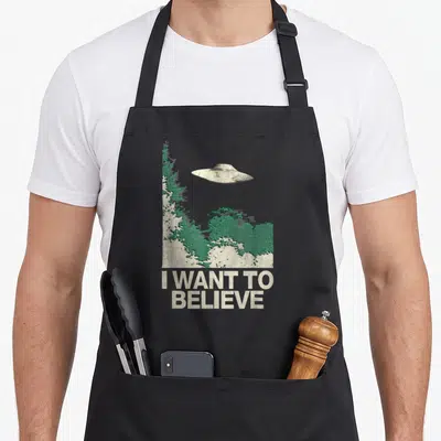 Tablier de cuisine noir avec illustration d'un ovni au-dessus d'une forêt verte et le texte I Want to Believe en bas.