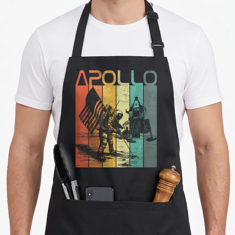 Tablier de cuisine noir avec motif Apollo rétro montrant un astronaute et le mot Apollo en lettres colorées verticales.