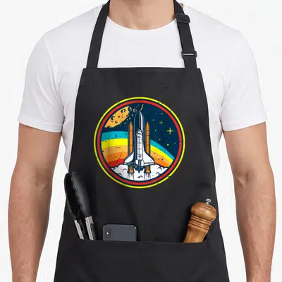 Tablier de cuisine noir avec écusson rond illustrant une navette spatiale blanche sur fond d'espace coloré et texturé.