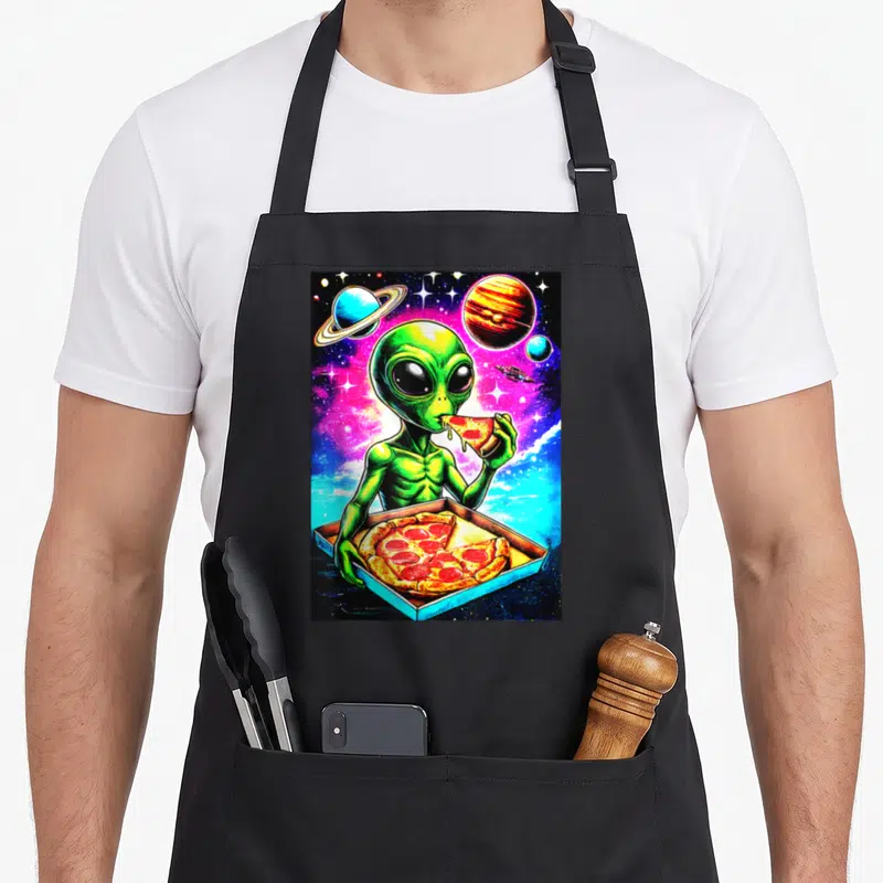 Tablier de cuisine noir avec un extraterrestre vert mangeant une pizza sur fond d'espace coloré.