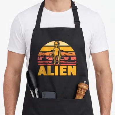 Tablier de cuisine noir avec illustration d'alien rétro sur fond de soleil couchant strié orange et rose.