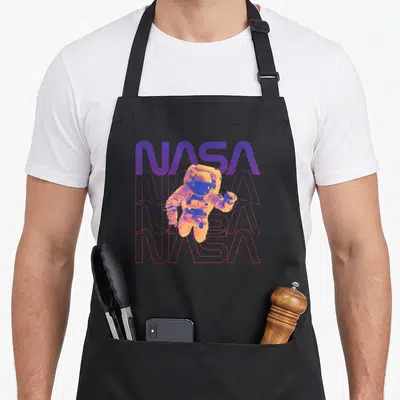 Tablier de cuisine noir avec un astronaute orange flottant devant des logos NASA superposés en violet et rose.