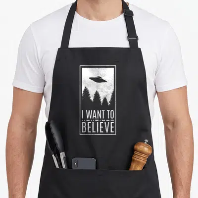 Tablier de cuisine noir avec illustration ufo forêt et texte I want to believe en blanc.