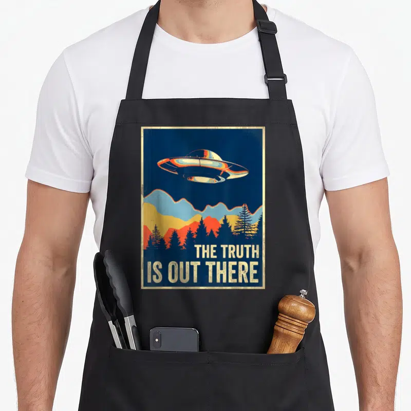 Tablier de cuisine noir avec illustration d'un ovni au-dessus de montagnes et forêts sous le texte The Truth Is Out There.