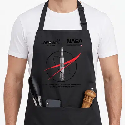 Tablier de cuisine noir avec illustration fusée Saturn V Apollo 11 et texte citation en anglais.