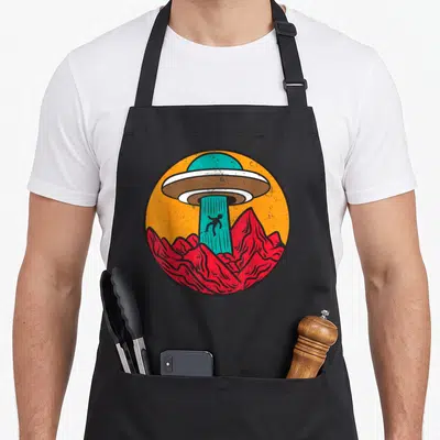 Tablier de cuisine noir avec illustration circulaire d'un ovni vert extrayant une silhouette humaine sur fond de montagnes rouges et ciel orange.