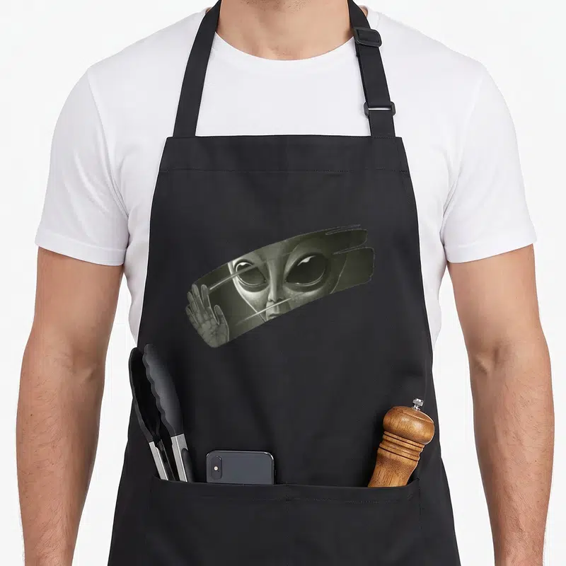 Tablier de cuisine noir avec illustration d'un alien aux grands yeux et d'une main tendue derrière une surface striée.
