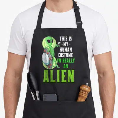 Tablier de cuisine noir avec extraterrestre vert tenant une soucoupe volante et texte humoristique sur le costume humain.