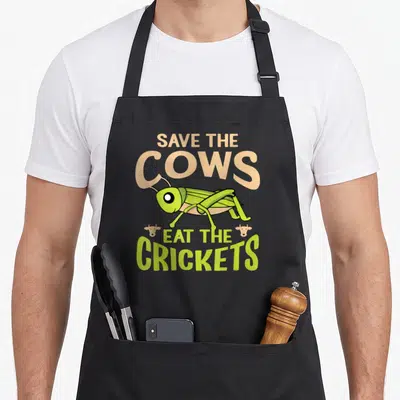 Tablier de cuisine noir avec illustration de criquet vert et texte Save The Cows Eat The Crickets en lettres colorées.