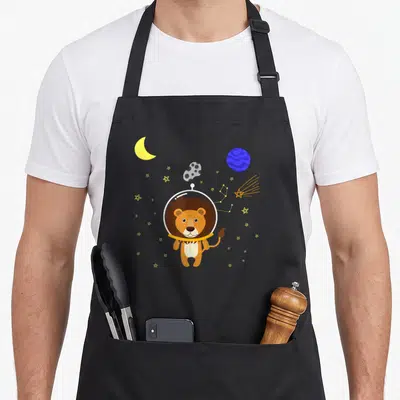 Tablier de cuisine noir avec un lion cosmonaute entouré de lune, planète et étoiles jaunes dans l'espace.