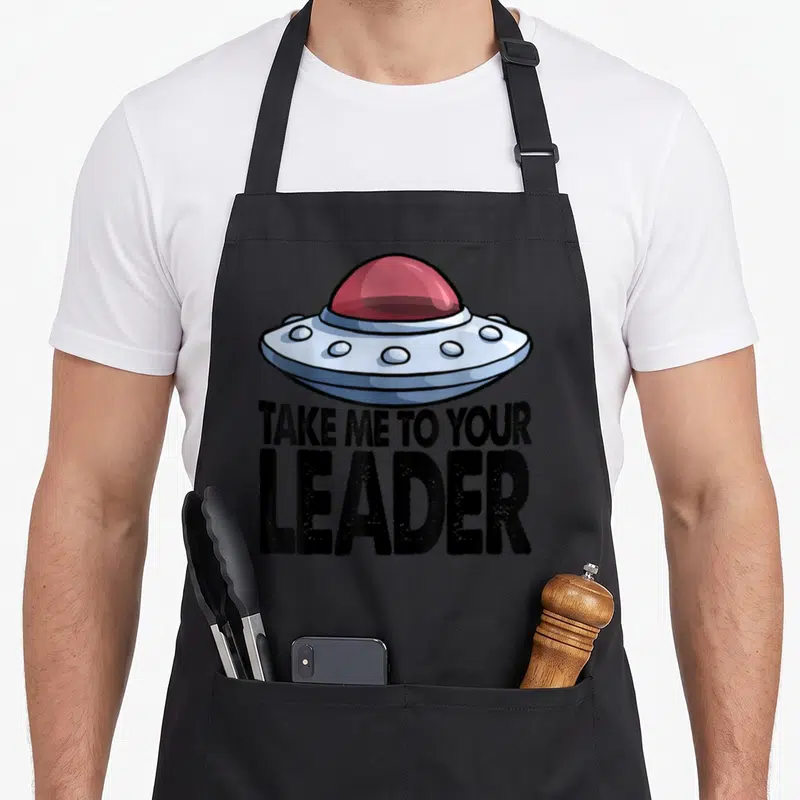 Tablier de cuisine noir avec illustration de vaisseau spatial et texte Take Me To Your Leader en noir.