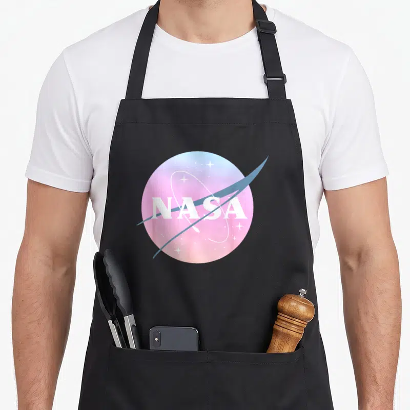 Tablier de cuisine noir avec un logo rond pastel rose et bleu portant l'inscription NASA et des étoiles blanches.