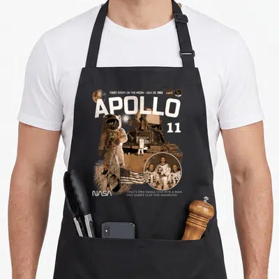 Tablier de cuisine noir avec illustration Apollo 11, astronaute, drapeau américain et logo NASA en tons sépia.