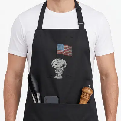 Tablier de cuisine noir avec Snoopy astronaute tenant un drapeau aux étoiles et bandes rouges.