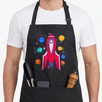 Tablier de cuisine noir avec une fusée rouge entourée de planètes colorées et d'étoiles dans l'espace.