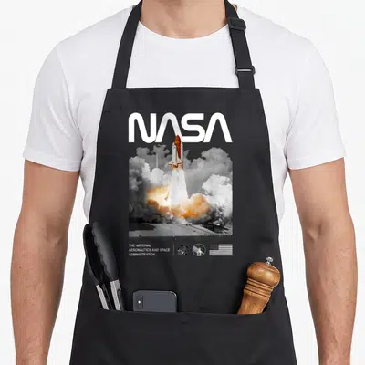Tablier de cuisine noir avec le logo NASA et une illustration colorée d'un lanceur spatial s'élevant dans des nuages de fumée.
