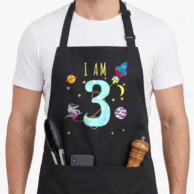 Tablier de cuisine noir avec un grand chiffre 3 bleu, une fusée et un astronaute colorés.