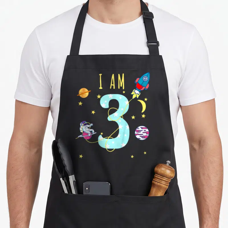 Tablier de cuisine noir avec un grand chiffre 3 bleu, une fusée et un astronaute colorés.