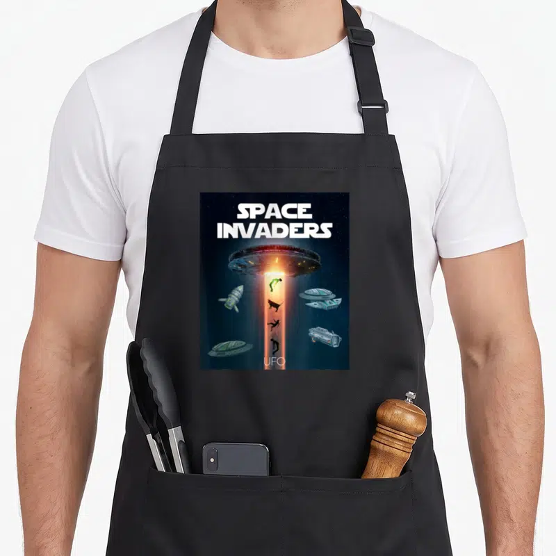 Tablier de cuisine noir avec illustration Space Invaders, ovni lumineux et vaisseaux spatiaux flottant dans l'espace.