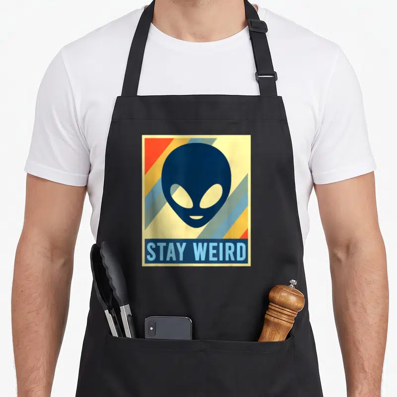 Tablier de cuisine noir avec illustration d'extraterrestre et texte Stay Weird sur fond rayé coloré.