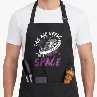 Tablier de cuisine noir avec une illustration de planète rose et le texte This Ace Needs Their Space en blanc et violet.