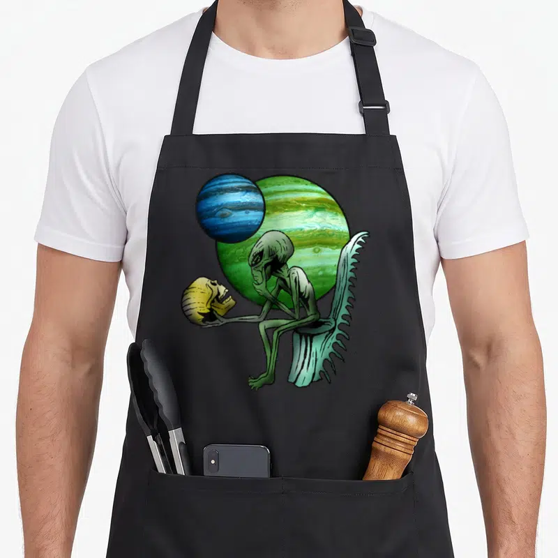 Tablier de cuisine noir avec un alien vert assis tenant une tête de mort devant deux planètes colorées.