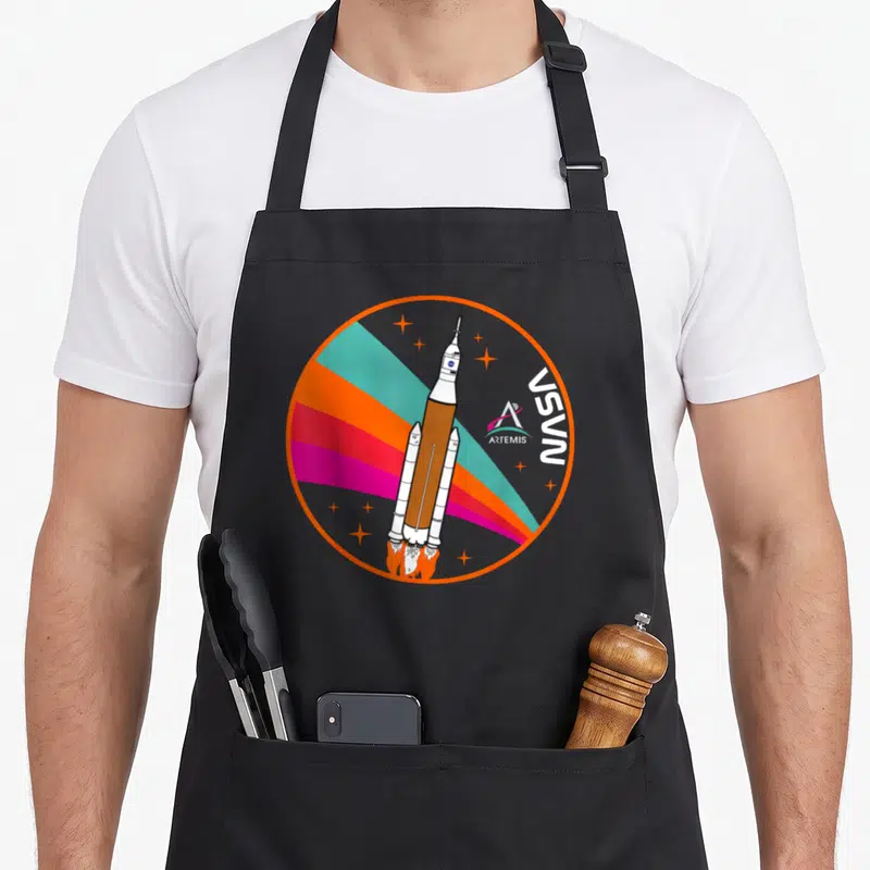 Tablier de cuisine noir avec illustration circulaire d'un lanceur SLS sur fond d'arc-en-ciel et logo Artemis.