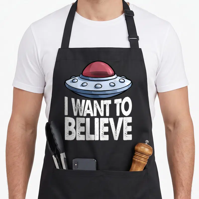 Tablier de cuisine noir avec illustration d'engin volant et texte I Want To Believe en blanc.