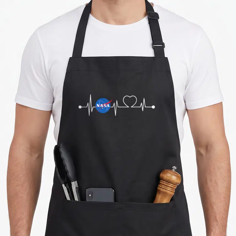 Tablier de cuisine noir avec une ligne de pulse blanche intégrant le logo NASA et un cœur stylisé au centre du motif imprimé.