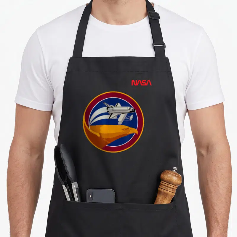 Tablier de cuisine noir avec l'emblème circulaire de la mission STS 51 G montrant une navette et un aigle doré.