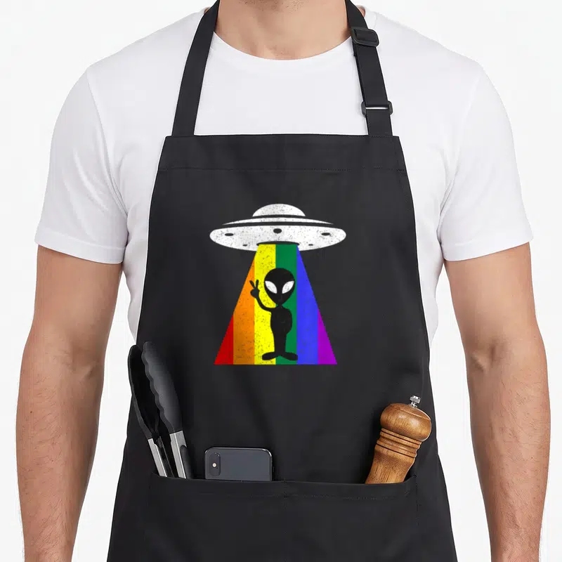 Tablier de cuisine noir avec un OVNI vintage projetant un rayon arc-en-ciel contenant un extraterrestre faisant la paix.