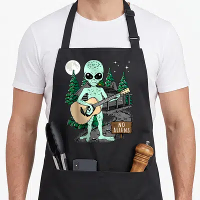 Tablier de cuisine noir avec un alien vert jouant de la guitare devant une cabane et un panneau NO ALIENS.
