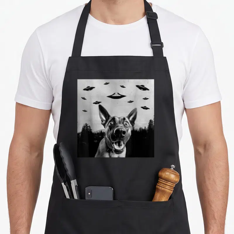 Tablier de cuisine noir avec illustration d'un Malinois souriant sous des vaisseaux volants en noir et blanc.