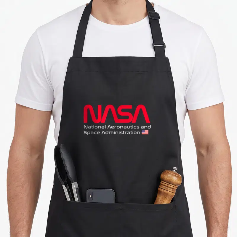 Tablier de cuisine noir avec le logo NASA en rouge et l'inscription blanche National Aeronautics and Space Administration accompagnée d'un petit drapeau américain.