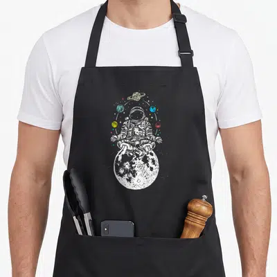 Tablier de cuisine noir avec illustration d'un cosmonaute méditant sur une lune texturée entourée de planètes et d'étoiles multicolores.
