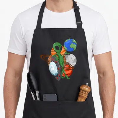 Tablier de cuisine noir avec extraterrestre vert posant sur un objet volant ovni devant des planètes du système solaire.