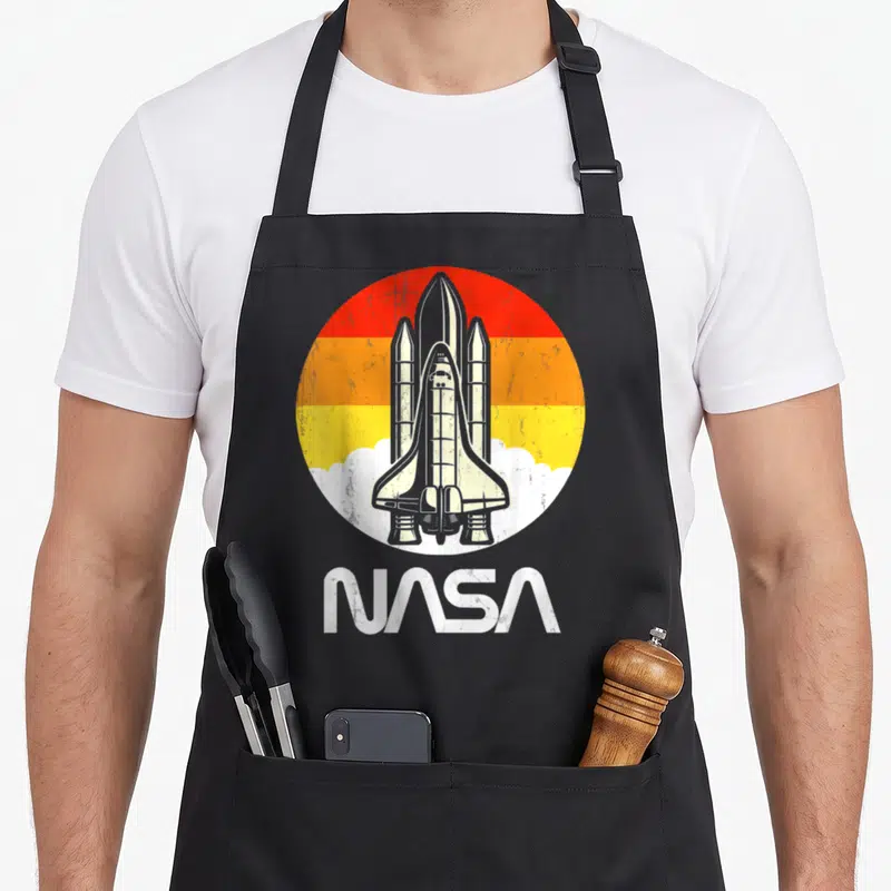 Tablier de cuisine noir avec illustration circulaire d'un lanceur spatial et nuages blancs sous un ciel orange dégradé.