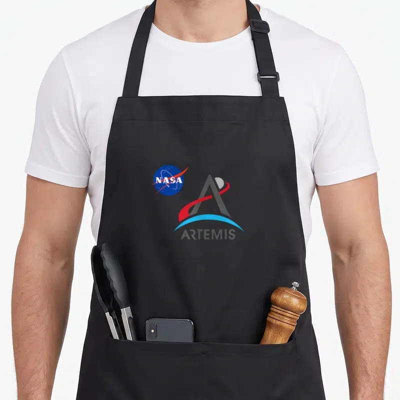 Tablier de cuisine noir avec le logo NASA et l'emblème stylisé de la mission Artemis imprimés au centre.