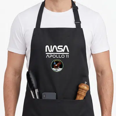 Tablier de cuisine noir avec le logo blanc NASA Apollo 11 et l'emblème circulaire de la mission lunaire au centre.