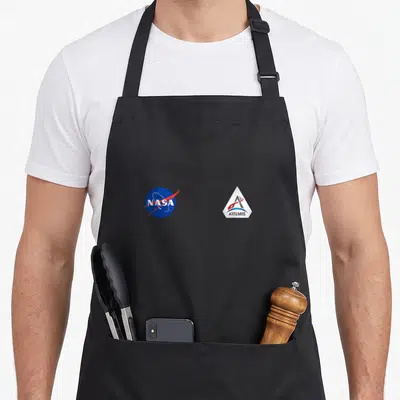 Tablier de cuisine noir avec les logos NASA et Artemis imprimés en couleur sur le devant.