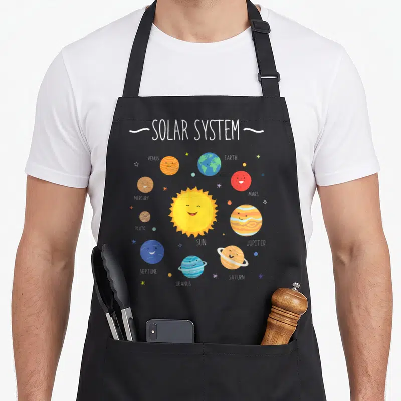 Tablier de cuisine noir avec illustration colorée du système solaire et des planètes aux visages souriants.