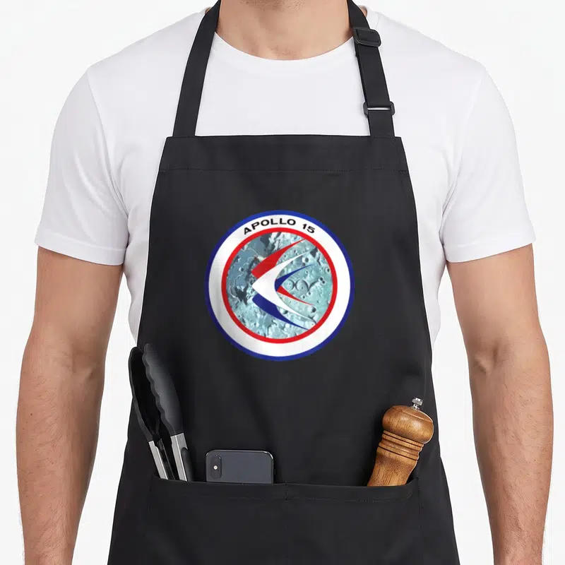 Tablier de cuisine noir avec l'emblème circulaire Apollo 15 sur fond de sol lunaire et motifs graphiques rouges et bleus.