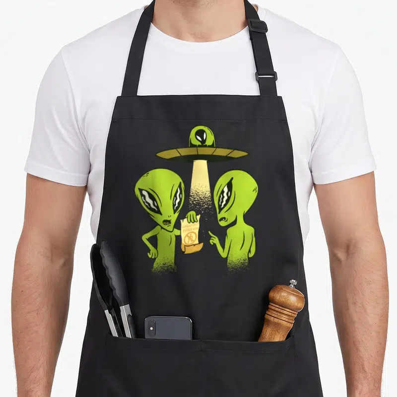 Tablier de cuisine noir avec deux extraterrestres verts et une soucoupe volante émettant un rayon lumineux sur un parchemin.