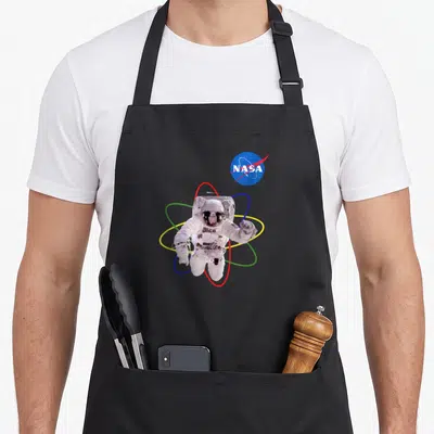 Tablier de cuisine noir avec un astronaute blanc flottant autour de cercles atomiques multicolores et le logo NASA bleu en haut à droite.