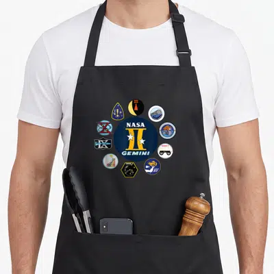Tablier de cuisine noir avec le logo NASA Gemini II et une couronne d'insignes de missions spatiales colorés sur fond bleu marine.