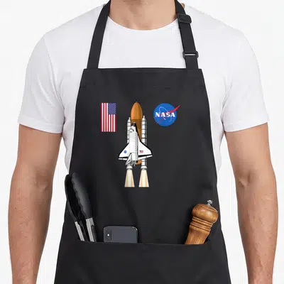 Tablier de cuisine noir avec une navette spatiale blanche, un drapeau américain et le logo bleu de la NASA imprimés sur le devant.