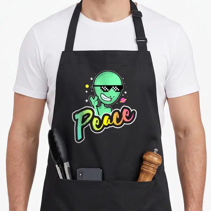 Tablier de cuisine noir avec un alien vert aux lunettes pixel et le mot Peace en lettres colorées.